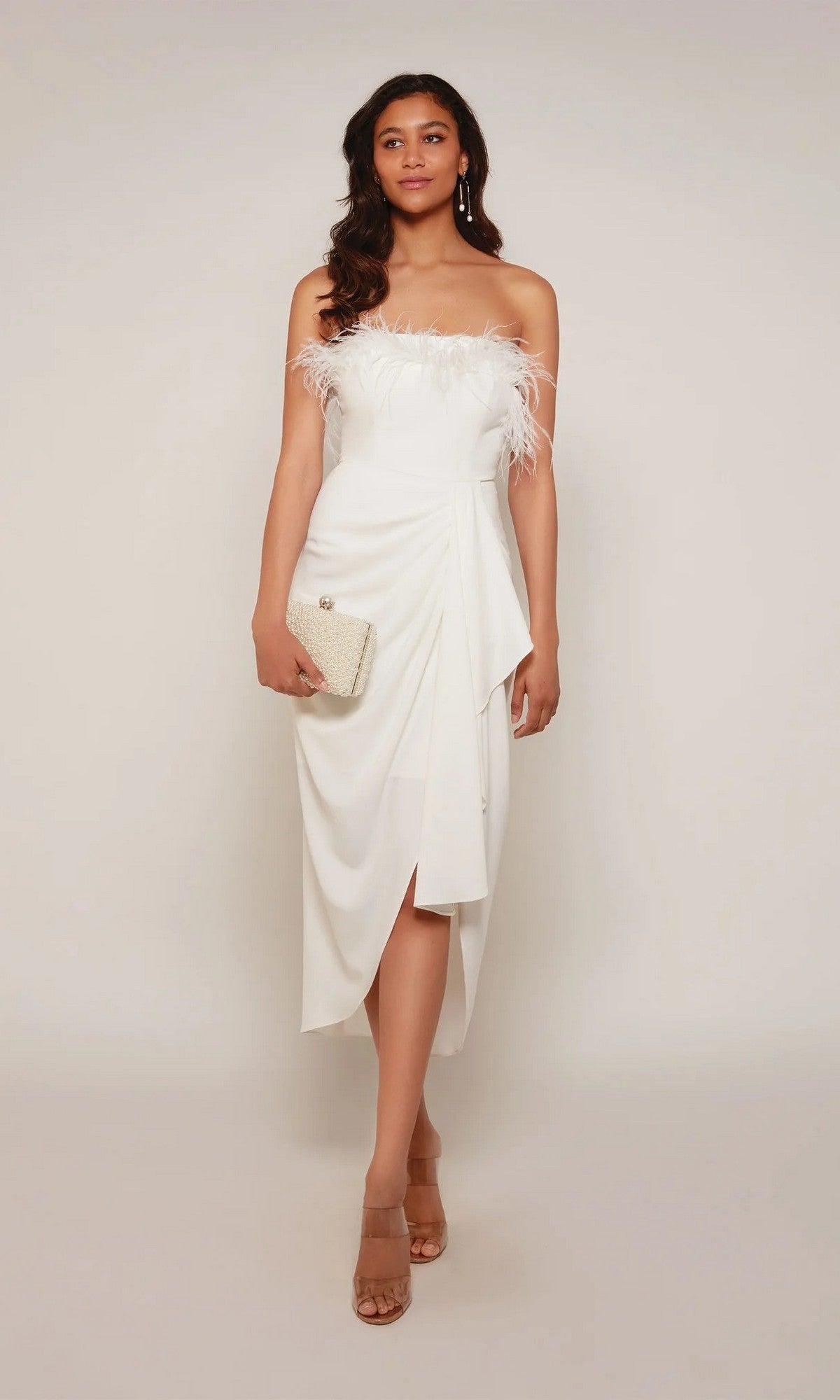 A woman in an ivory Alyce 70034 knee-length semi-formal dress with feather accents holds a small clutch and stands in sandals against a plain background.